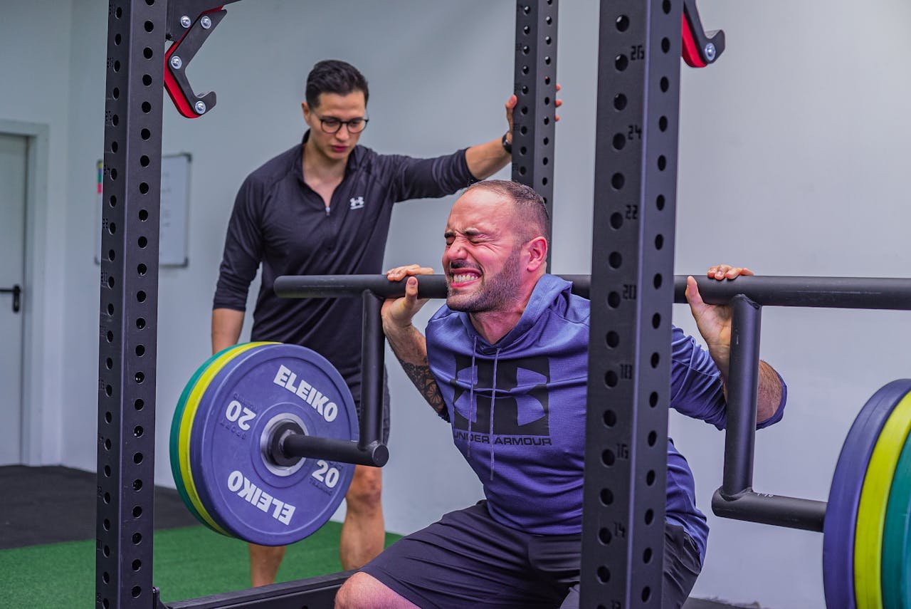 A man intensely performs a squat exercise with a barbell while his coach guides him in the gym.